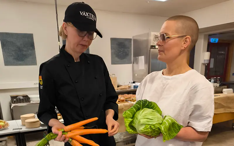 Två kvinnor i ett kök. I händerna har de morötter och grönsallad. 