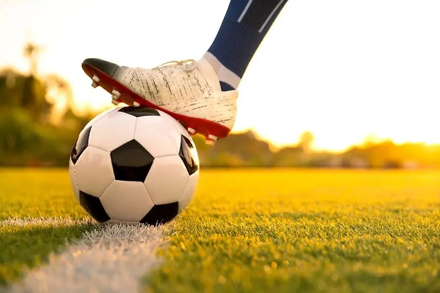 En fot med en fotbollssko håller foten på en fotboll. Fotbollen ligger ner på ett vitt streck på en fotbollsplan.